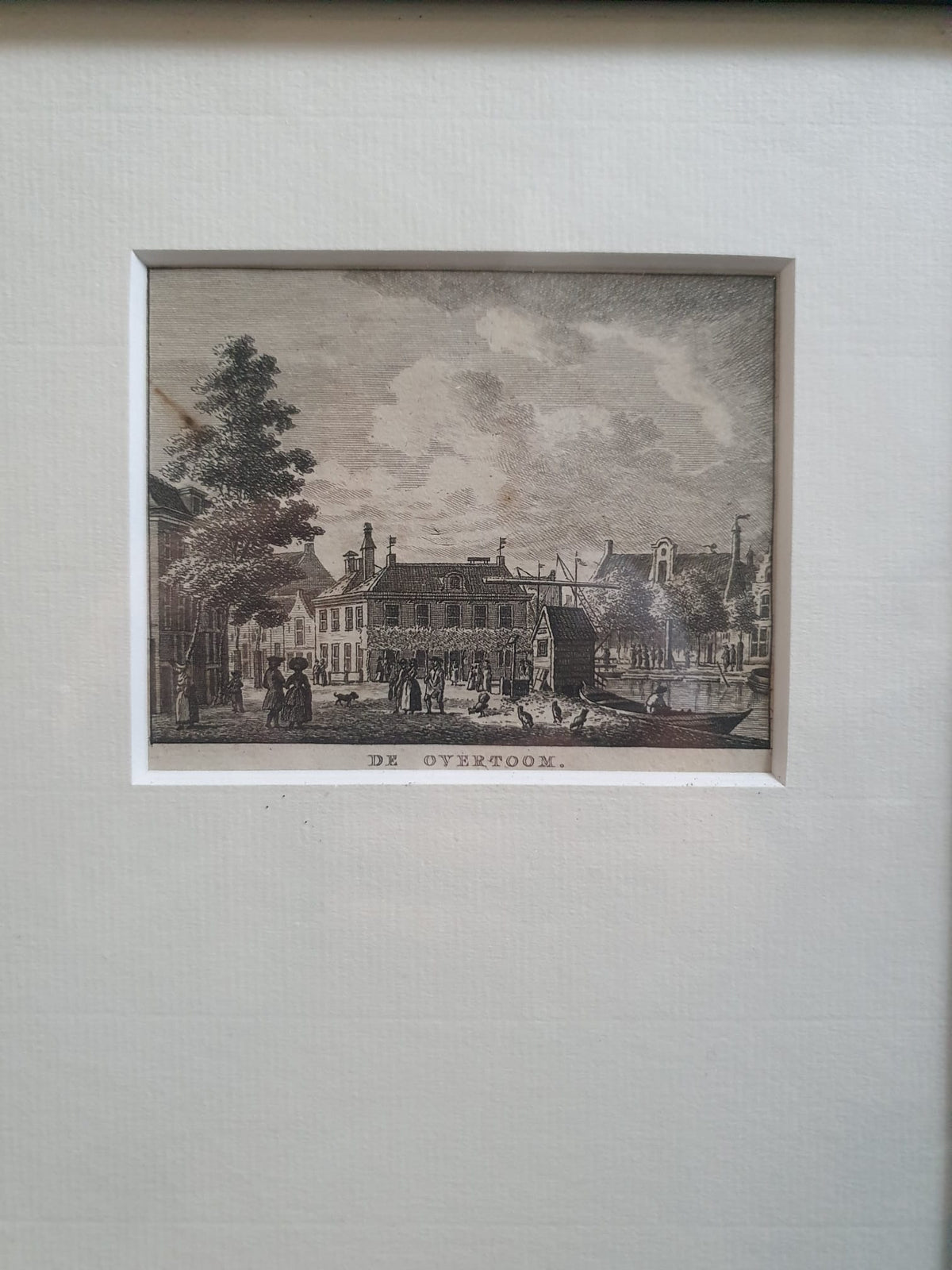 Ingelijste kopergravure met stadsbeeld aan water en mensen, opschrift onderaan